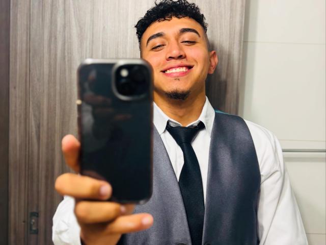 DakotaTheo with curly black hair, smiling, wearing a white shirt, black tie, and gray sash, holding a black phone in front of him, selfie in a bathroom.