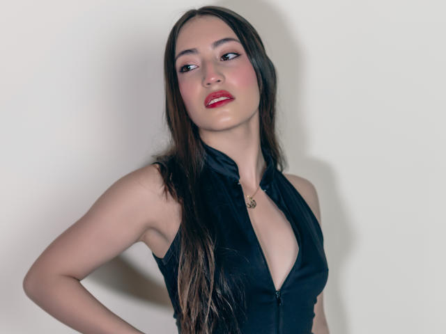 AnnFisher has long brown hair, red lipstick, and a black halter top with a deep cleavage, showing her small tits and a gold necklace. She looks sideways with a smug expression.