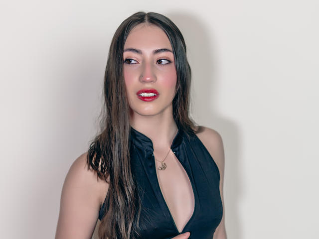 AnnFisher with long brown hair, red lipstick, and black halter top, showing a deep cleavage, looking to the side, wearing a small necklace.