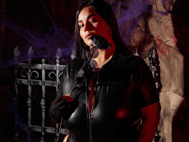NohayMuller with dark hair, black leather outfit, and black gloves, holding a black feather in her mouth, standing in front of a metal fence, smoky purple background.
