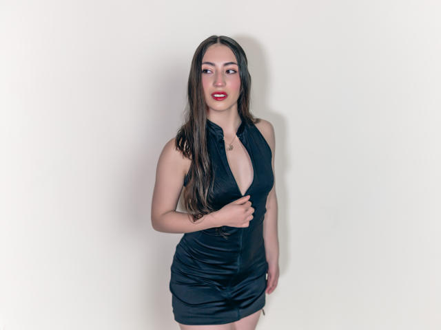 AnnFisher with long brown hair, red lipstick, and black dress, exposing her big tits and cleavage, standing against a white wall, holding dress, looking at camera.
