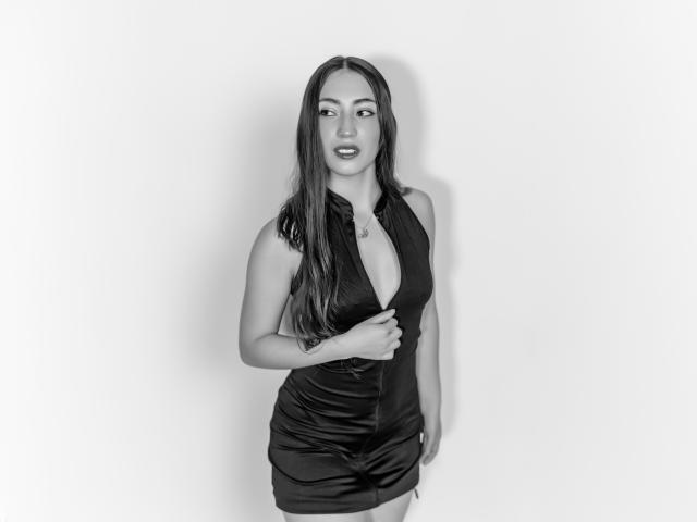 AnnFisher in a black, deep V-neck, sleeveless dress, exposing her tits, long dark hair, tattoo on her right forearm, white background, standing, looking to the side.