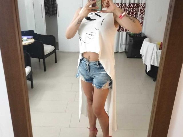 SammyDesire has a white sleeveless crop top with a face print, frayed denim shorts, and a long white drape. She's holding a phone, showing tattoos on her arms.