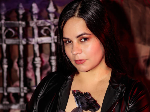 NohayMuller with dark straight hair, wearing a black leather jacket, has pinkish-red eyeshadow, a slight blush, and a black rose accessory near her tits.