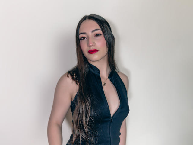 Ann Fisher hat langes, braunes Haar, roten Lippenstift und ein schwarzes Top ohne Ärmel mit tiefem Ausschnitt. Sie steht vor einer schlichten Wand und schaut in die Kamera..