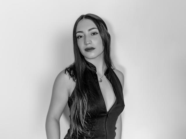 Ann Fisher, avec ses longs cheveux noirs, portant un haut noir sans manches avec un décolleté en V plongeant, dévoilant ses seins, debout devant un fond uni, sur une photo en noir et blanc..