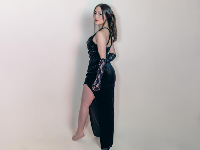 Ann Fisher vestida con un vestido de terciopelo negro con mangas de encaje, mostrando su trasero con una abertura alta, de pie descalza, mirando por encima del hombro con una ligera sonrisa..