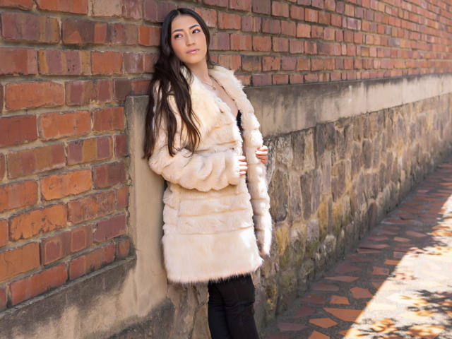 AnnFisher with long brown hair, wearing a fluffy beige fur coat, black pants, and black boots, leaning against a brick wall, smiling slightly, hands in coat pockets.