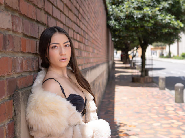 AnnFisher with long brown hair, wearing a fluffy white and black fur coat, leaning against a red brick wall, showing small boobs, and looking at the camera.