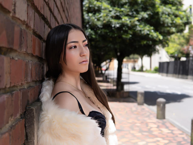 AnnFisher with long brown hair, pale skin, wearing a black bra and white furry coat, leaning against a brick wall, looking to the side, sunny day, street in background.