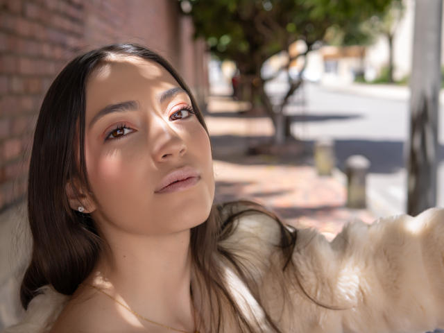 AnnFisher has smooth brown skin, long dark brown hair, and brown eyes. She wears a fluffy white coat, and her soft smile shows pink lipstick. Her small tits are partially visible.