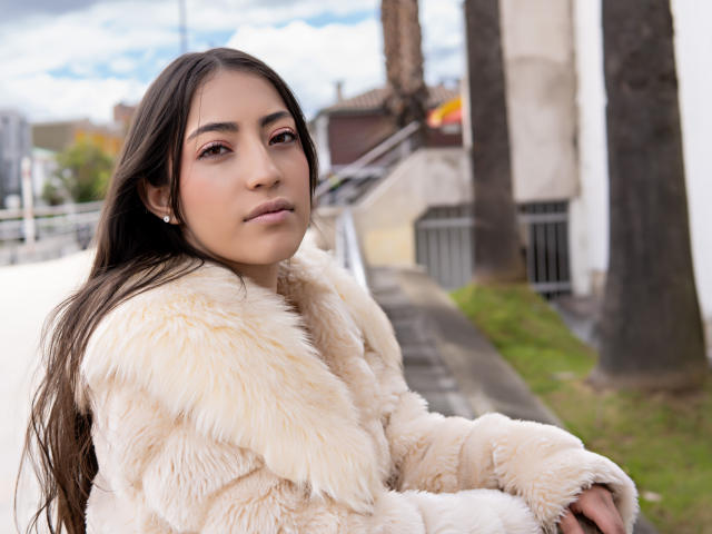AnnFisher with long brown hair, wearing a fluffy beige fur coat, has her ass slightly turned, looking over her shoulder with a subtle smile, standing outdoors.