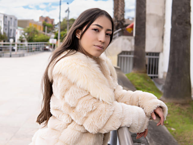AnnFisher with long brown hair, wearing a fluffy beige fur coat, standing by a metal railing, looking over her shoulder with a slight smile, holding the railing with her hands.