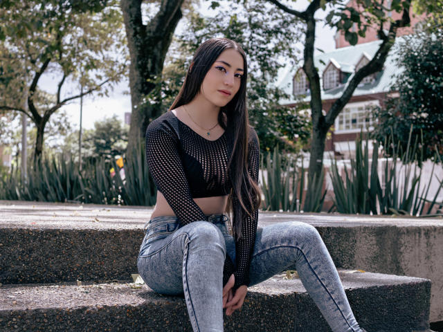 Ann Fisher, con cabello largo y oscuro, vestida con una blusa de malla negra y jeans azul claro, sentada en escalones de concreto, mirando a la cámara, con las manos sobre las rodillas, al aire libre, durante el día..