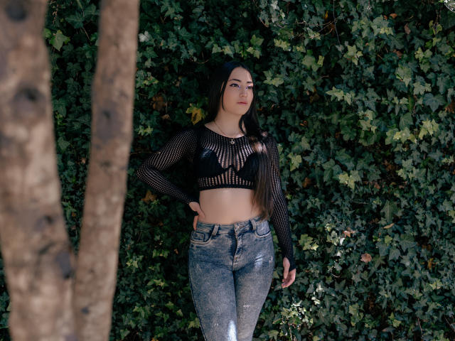 AnnFisher with black mesh top showing her small tits, wearing high-waisted, shiny blue jeans, standing against a leafy wall with her hand on her hip.