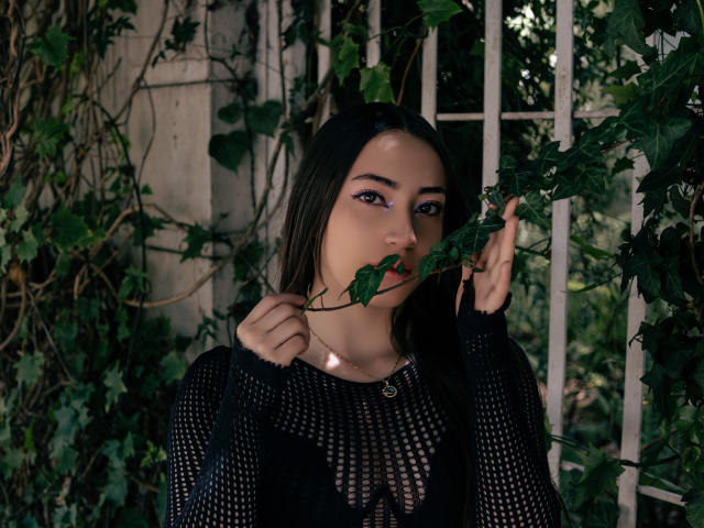 AnnFisher with dark hair, purple eyeliner, black mesh top, holding green leaf in mouth, looking at camera, surrounded by vine leaves, in a slightly dark, moody setting.
