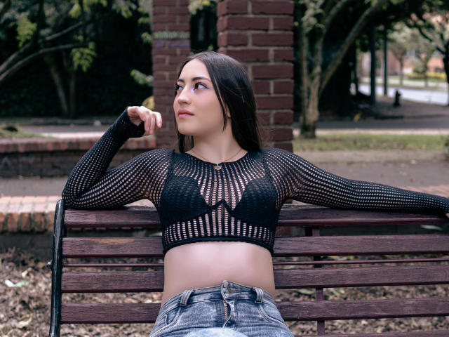 AnnFisher with medium tits, wearing a black mesh crop top showing her black bra, and blue jeans, sitting on a wooden bench in a park, looking up with a slight smile.