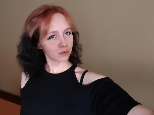 AnnaHypnosis with pink and black dyed hair, off-shoulder black top, blue eyes, and subtle makeup, looking slightly to the side, showing a bit of her bare shoulder.