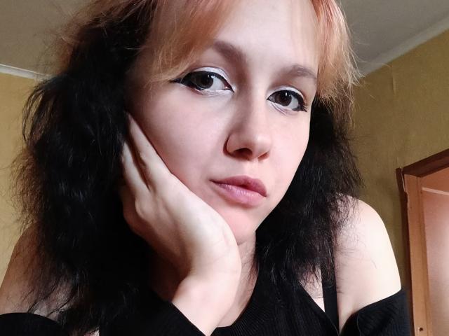 AnnaHypnosis has black and blonde curly hair, pale skin, and black eyeliner. She rests her hand on her cheek, looking slightly down with a neutral expression.