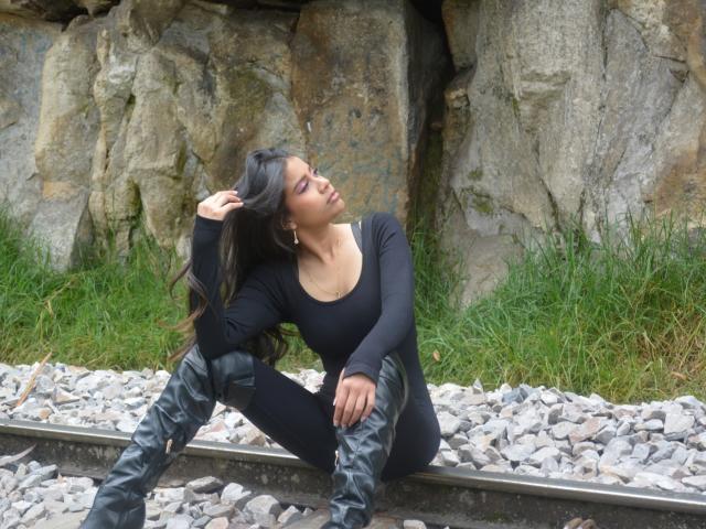 Adaallen with long black hair, wearing a black long-sleeve top and camo knee boots, sitting on railroad tracks, hand in hair, eyes closed, leaning back with a relaxed expression.