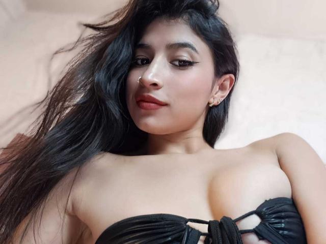 NaiaSanders with long black hair, nose ring, and eyeliner, wearing a black strapless top, showing her tits, lying down, looking at the camera with a slight smile.