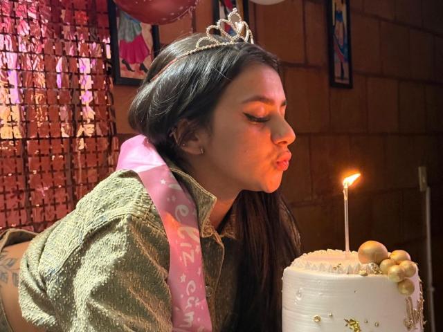 AlessiaFerry with long black hair, a glittery green shirt, and a tiara, pouting while blowing out a candle on a white cake with gold decorations.
