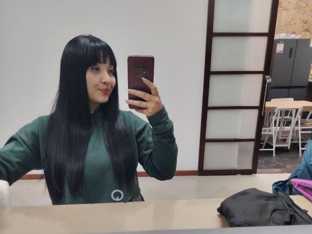LunaBellaHotty with long black hair, green sweater, holding phone, taking mirror selfie, smiling, boobs slightly visible, mirror reflection, phone case is maroon, clothes on table.