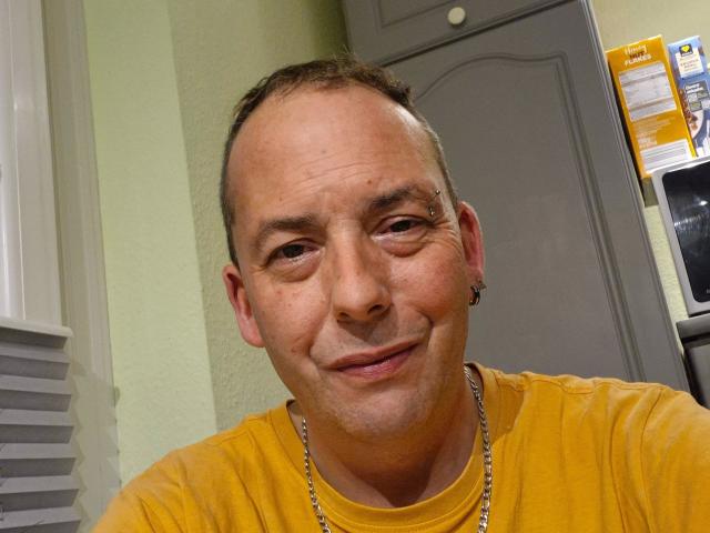 Rocketman-hot, bald with short brown hair, smiling, wearing a yellow shirt, silver chain, small nose ring, slightly wrinkled face, greenish wall behind him, looking casual and relaxed.
