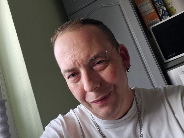 A close-up of Rocketman-hot, middle-aged man with a balding head and short brown hair, wearing a white t-shirt and a silver chain necklace. He has a small metal stud in his right eyebrow and a black ear cuff. His expression is slightly smiling with a relaxed, slightly wrinkled face. Natural light from the left. Background includes a grey door and a shelf with a black TV on the right. Simple, everyday setting.