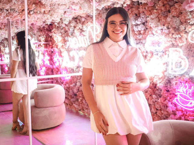 CamilaSofiia with long dark hair, smiling, wearing a pink and white checkered vest over a white dress, standing in a pink flower-covered room, reflection in mirror.