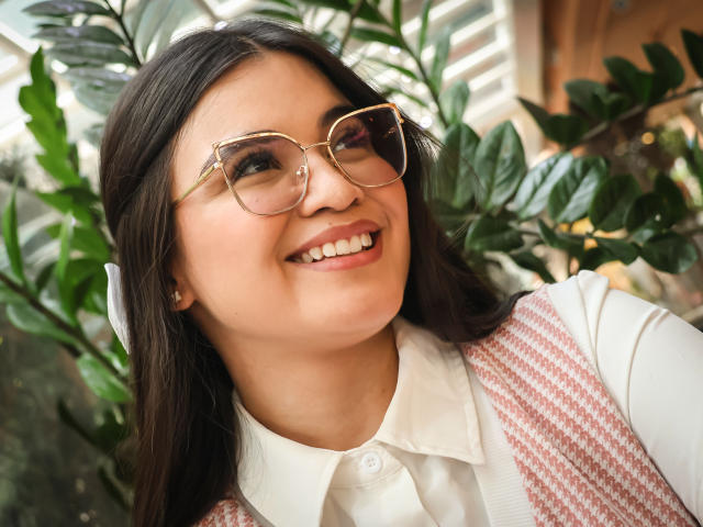 CamilaSofiia with glasses, smiling, brown hair, white shirt, pink houndstooth vest, looking up, green plant leaves in background, soft natural light, cute, attractive, slightly chubby face.