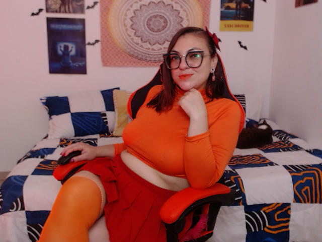 LunaRausch, with red pigtails, orange glasses, and orange top, sits in a chair, wearing a red skirt and orange thigh-highs, showing her white-belly and orange-boobs.
