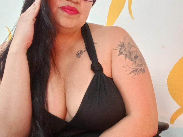 Ginebra Vargas, con cabello negro largo, vestida con un top negro de tirantes que muestra un busto grande, tatuaje en el brazo derecho, lápiz labial rojo, gafas negras, tocándose la cara..