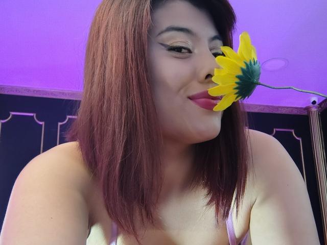 Noemi Thornee, con i capelli castano-rossicci, indossa un reggiseno rosa e sorride a un girasole giallo che le copre il naso, con uno sfondo illuminato di viola. Il suo eyeliner è ben definito, le labbra sono rosa..