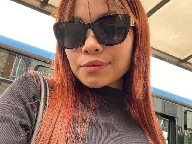 VannesaSaenz with red hair, wearing black sunglasses, and a black turtleneck, showing a slight pout, holding a black strap, standing near a bus.