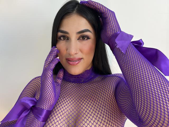 AlanaHarris in purple fishnet lingerie, with matching gloves and ribbons, has her hands in her long black hair, showing off her sexy, curvy tits and confident smile.