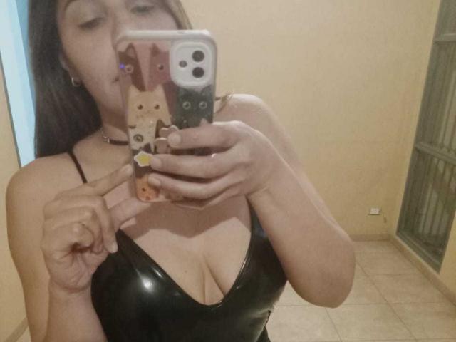 NatashaN with tan skin, wearing a black leather bra, taking a mirror selfie with a phone case featuring a cat face, pointing at her tits with a finger.