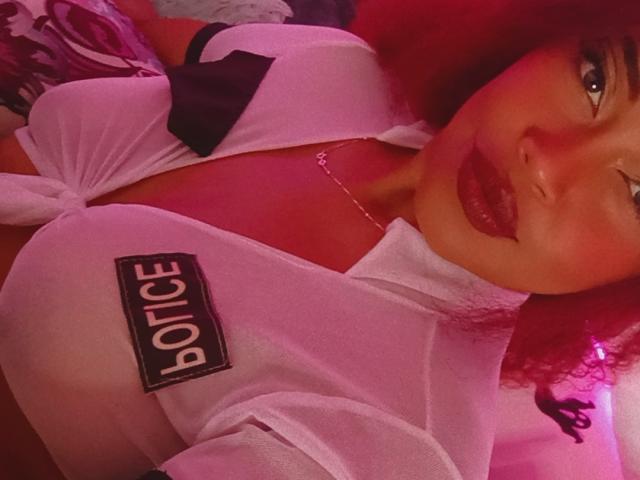 DalilaHeinz met rood krullend haar, knipend, draagt een doorschijnend wit topje dat onder haar grote borsten is vastgebonden, een zwarte strik en rode lippenstift, een delicate ketting, en er is roze verlichting..