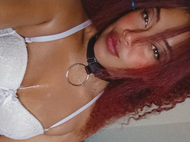 DalilaHeinz with red curly hair, white lace bra, and black collar with metal ring, glistening skin, smiling, showing small to medium tits, and brown eyes.