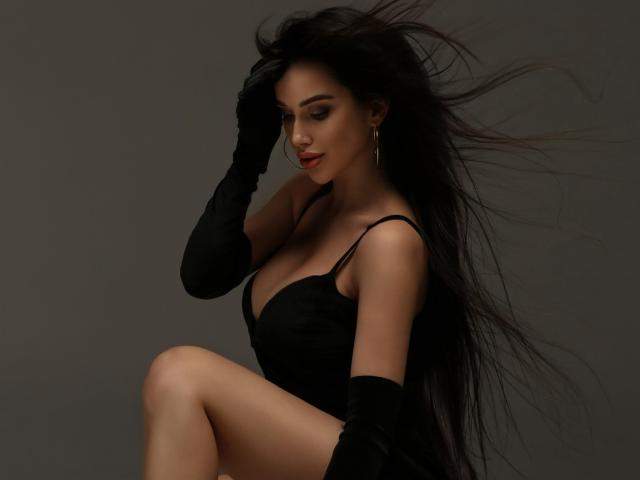 Tayor69 with long black hair, wearing a black lace dress and gloves, showing her tits, sitting with one leg up, looking down, dark gray background.