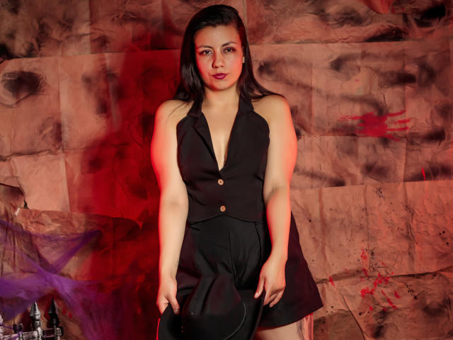 MargarettCooper with medium tits, black sleeveless vest, and black shorts, standing, red and purple smoke, dark hair, slight smile, fair skin, brown eyes, in a dimly lit room.