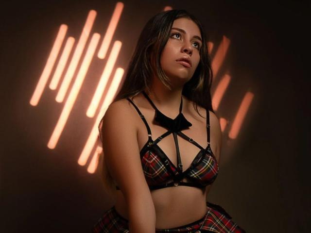 CloeSweetsX stands center in a dark, dimly lit space with warm, vertical light streaks in the background. She has long, straight brown hair and a slightly pensive expression. She wears a red plaid bra with black straps and a small black bow in the center, paired with a matching skirt. Her skin tone is light, and she has a youthful, attractive appearance. The light streaks create a modern, almost futuristic aesthetic. Her outfit is bold and provocative, with a focus on the plaid pattern and black accents. The composition is balanced, with CloeSweetsX slightly off-center to the right.