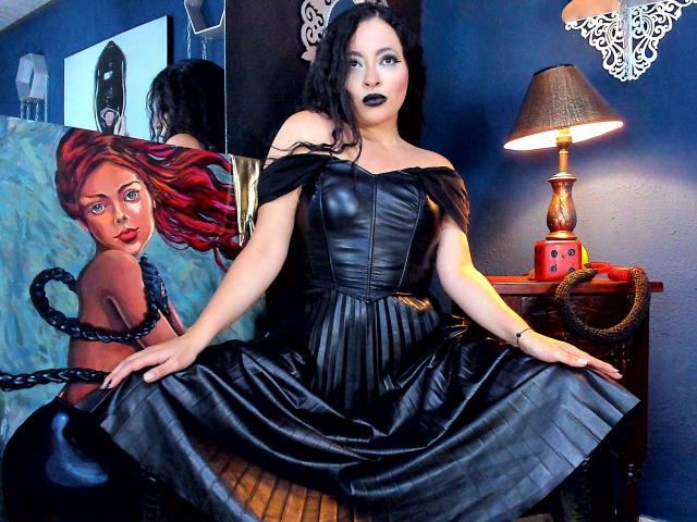 LunaTikka with dark lipstick, black off-shoulder leather dress, and black skirt, sitting on wooden chair, black chain in hand, red lamp on right, painting of red-haired woman behind.
