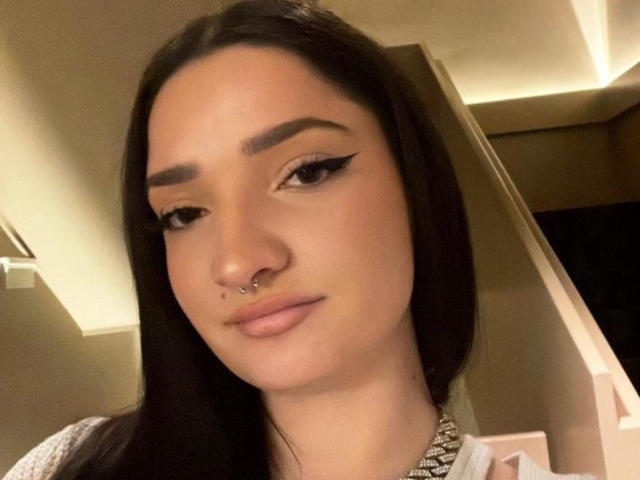 VelvetSelini with long dark hair, thick eyebrows, winged eyeliner, nose ring, and a subtle smile, wearing a sparkly necklace, in a softly lit indoor setting.