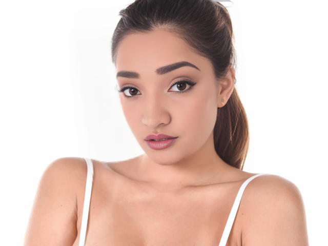 VelvetSelini has medium brown skin, dark brown eyes, and full pink lips. She's wearing a white spaghetti strap top, with her brown hair in a ponytail.