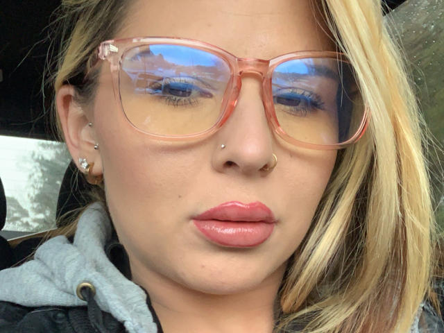 MonyBelle with pink glasses, blonde hair, nose ring, piercings, glossy pink lips, and reflective eyewear showing a cloudy sky. Wearing a gray hoodie.