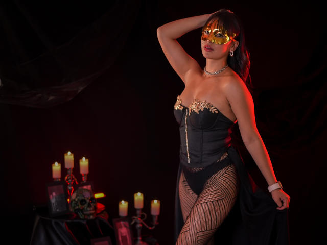 SharaaMiller with a curvy ass, wearing a black corset, fishnet stockings, and a gold mask, posing against a dark background with candles, showing off her tits and a black cape.