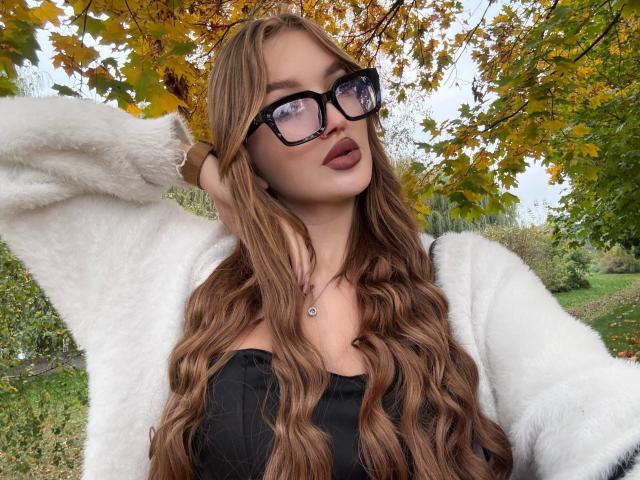 KlarnetHot has wavy brown hair, wears black glasses, a white fuzzy cardigan, and a black top, with pouty, dark-red lipstick, and a necklace with a small heart.