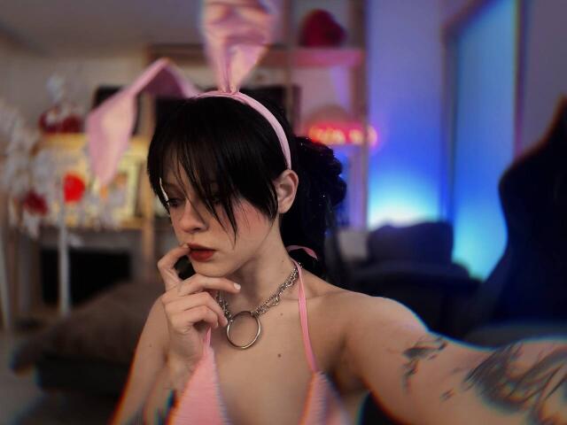SimaWendy with pink bunny ears, black hair, red lipstick, chain collar, tattoos, and a pink halter top, thinking with finger on chin, in a blurred room.