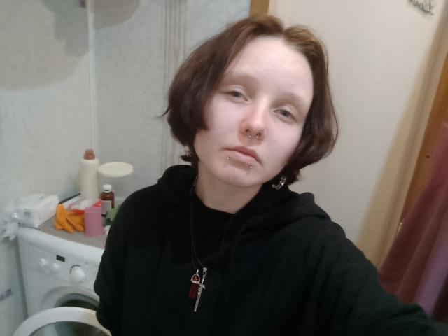 OliviaFlex, a young woman with pale skin and short brown hair, stands in a bathroom. She wears a black hoodie and a necklace with a red pendant and key. She has a nose ring and a lip piercing. The photo is slightly blurry. The background includes a white washing machine, a pink box, and a beige wall. The overall composition is casual and unrefined.